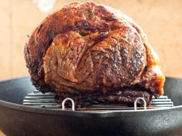 Browned prime rib roast on elevated rack in a cast iron pan on a wood cutting board. Close up.