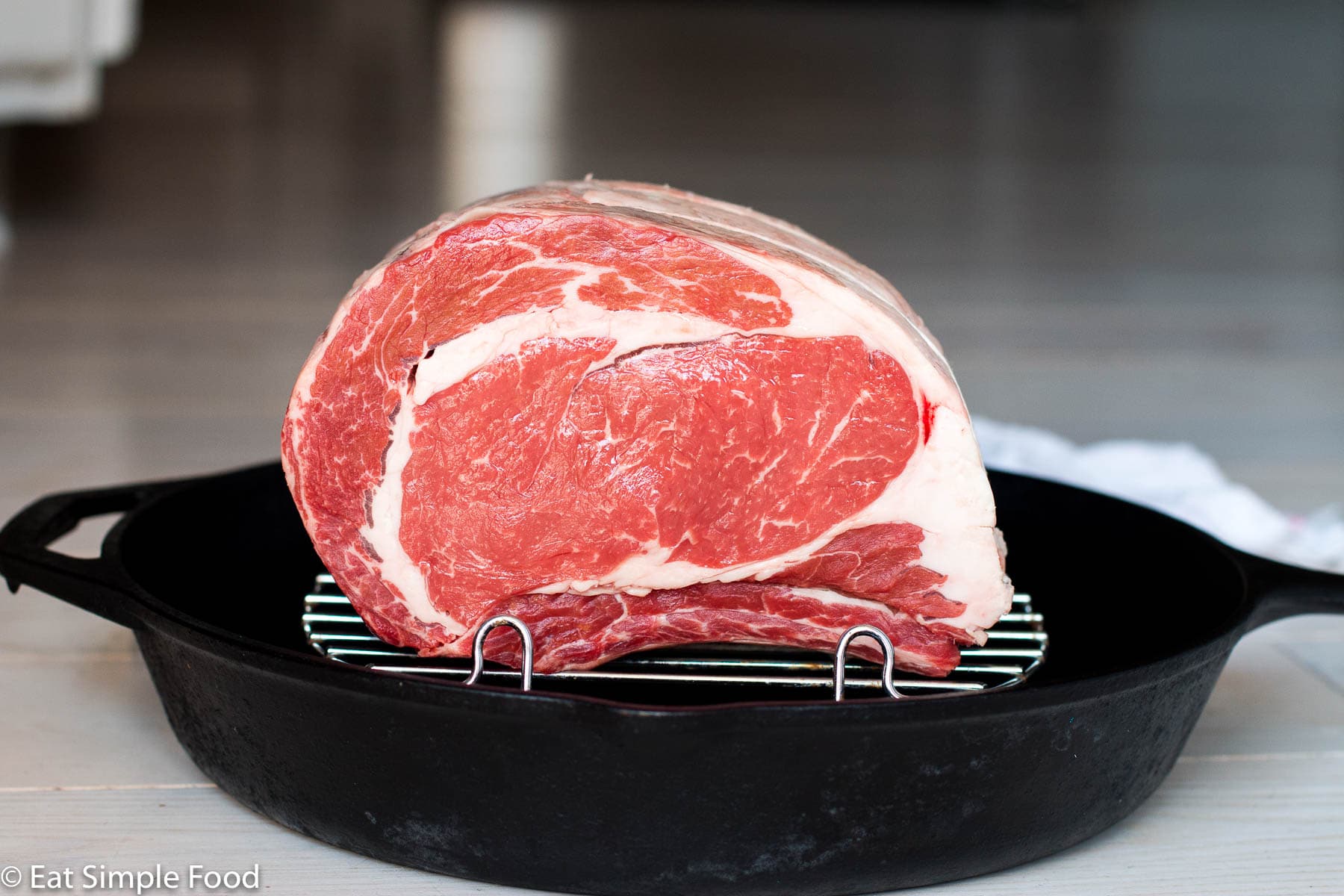 Raw prime rib roast on elevated rack in a cast iron pan on a white table. Side view.