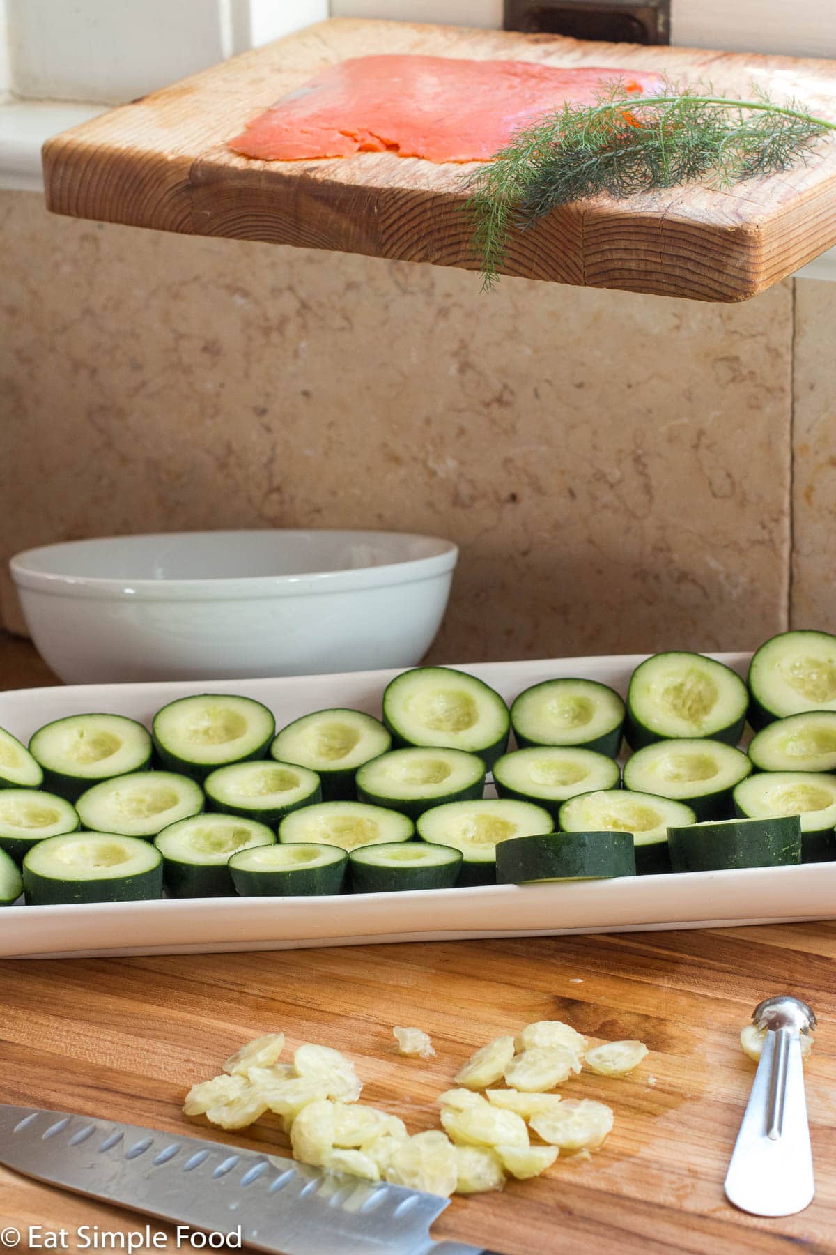 Two wood cutting boards. One on a windowsill with a flat piece of smoked salmon and dill sprigs. The other on a cutting board with a plate of cucumber rounds with a white bowl in the background and a knife in the front.