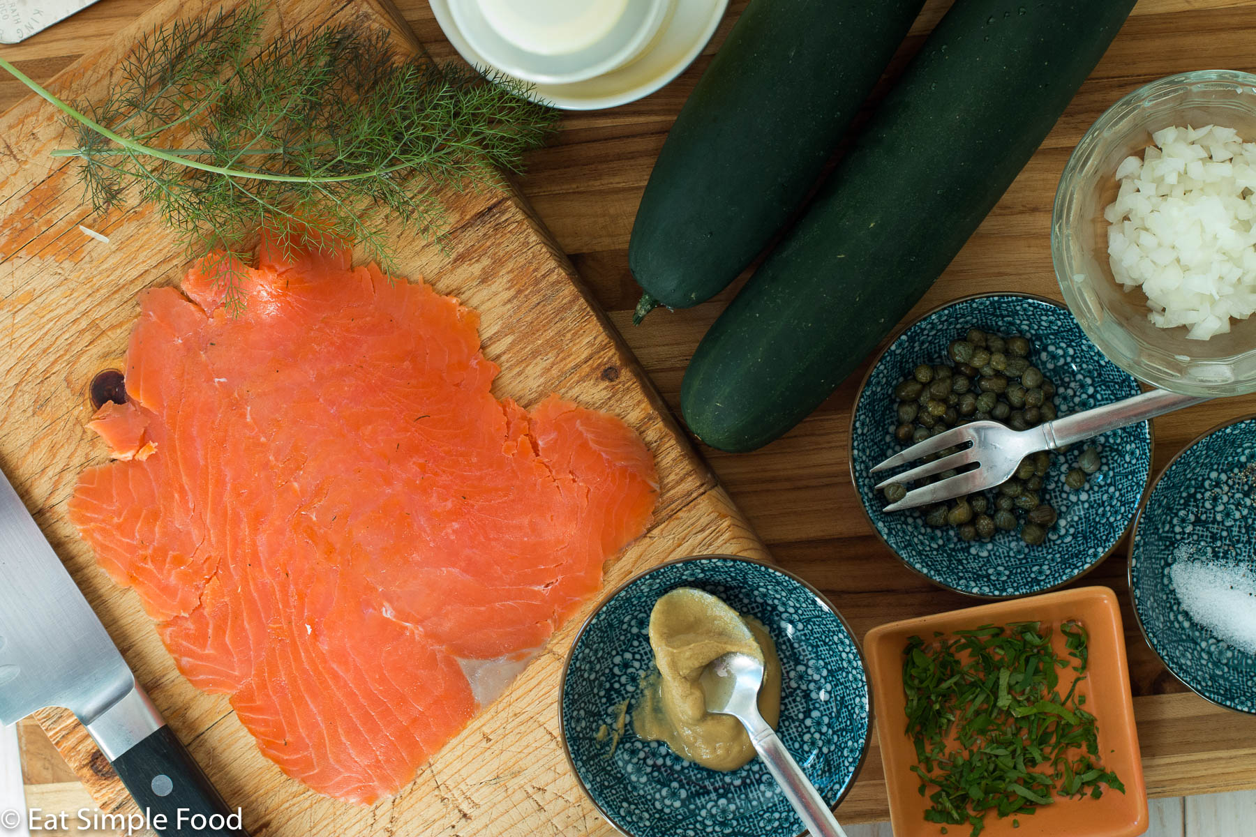 Ingredients on wood cutting board. Top down view of smoked salmon, whole cucumbers, bowl of capers, bowl of diced onions, dill sprigs, bowl of mustard, bowl of chopped parsley.