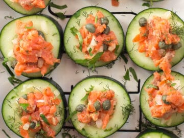 White rectangle plate of smoked salmon cucumber rounds with capers, onions, and chopped dill and parsley. Top view.