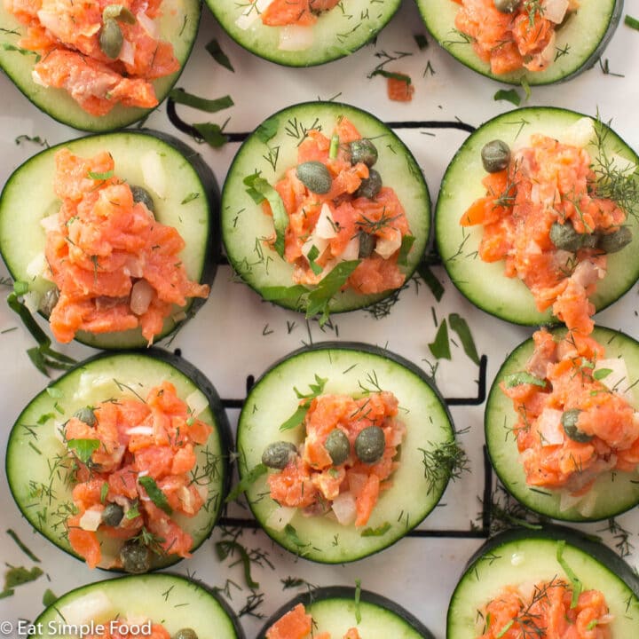 White rectangle plate of smoked salmon cucumber rounds with capers, onions, and chopped dill and parsley. Top view.
