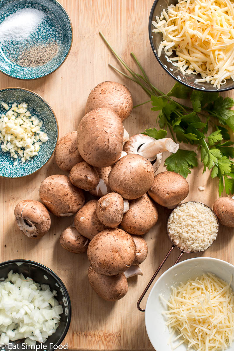 Ingredients on wood cutting board: round bows of salt and pepper, minced garlic, shredded long cheese, grated Parmesan cheese, diced onions, and bread crumbs, whole mushrooms and sprigs of parsley.