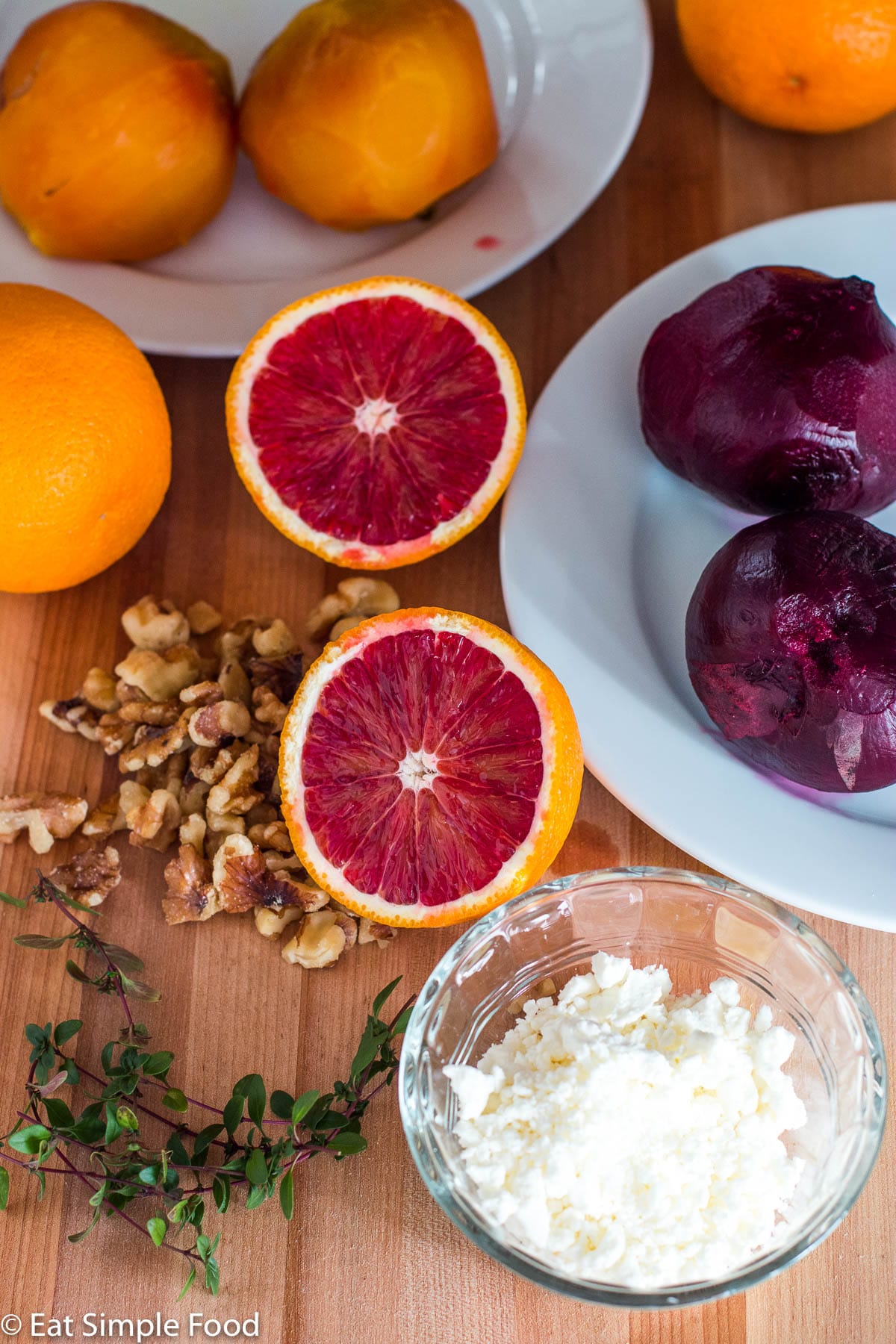 Ingredients on a wood cutting board: cooked whole red beets, cooked whole yellow beets, whole oranges, walnut pieces, small cup of goat cheese crumbles, and sprigs of thyme.
