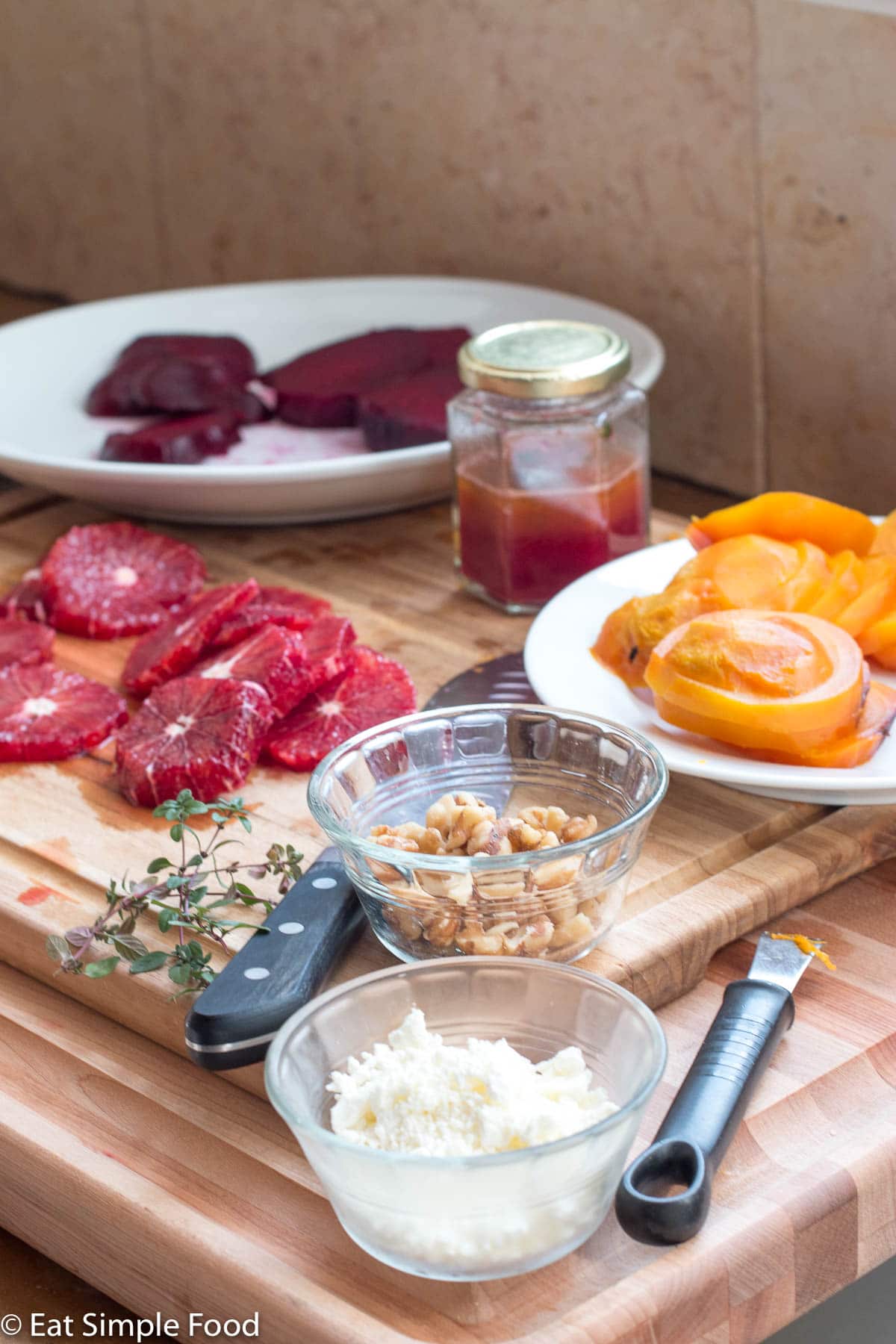 Ingredients on a wood cutting board: cooked sliced red beets, cooked sliced yellow beets, peeled sliced oranges, small cup of walnut pieces, small cup of goat cheese crumbles, sprigs of thyme, and a chef's knife.