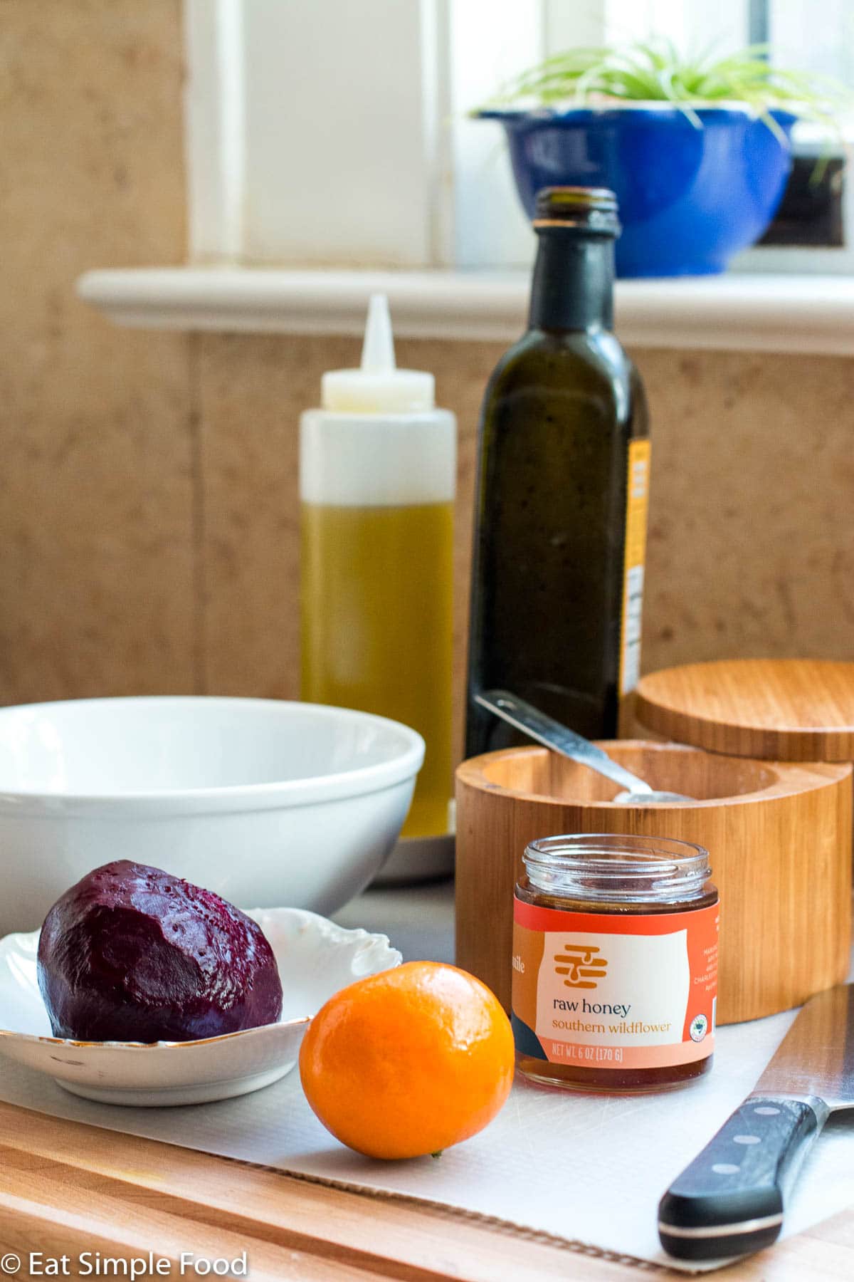 Ingredients on a wood cutting board in front of a windowsill: cooked and peeled whole red beet, orange, jar of honey, bottle of olive oil, bottle of balsamic, wood bowl of salt, white bowl, and chef's knife.