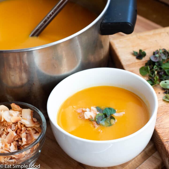 Side view of white bowl of orange yellow butternut squash soup with a spoon on the side and toasted coconut flakes and mint on the side. Stainless steel pot of soup in background with a ladle.