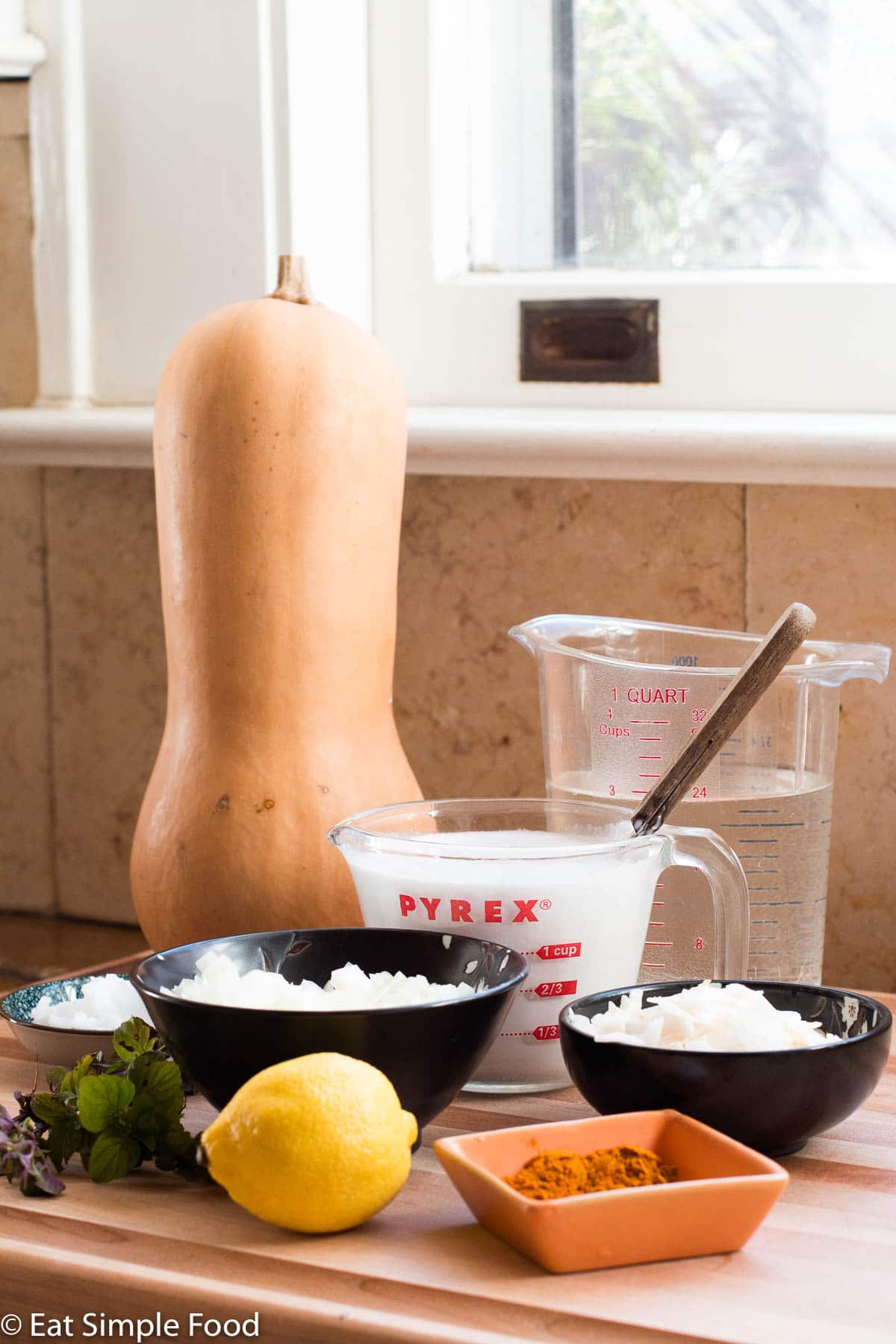 Ingredients on wood cutting board in front of a window: large butternut squash standing up, measuring cup full of coconut milk, measuring cup of 3 cups water, small bowl of curry, diced onions, coconut oil, coconut flakes, and one whole lemon. Mint on the side.