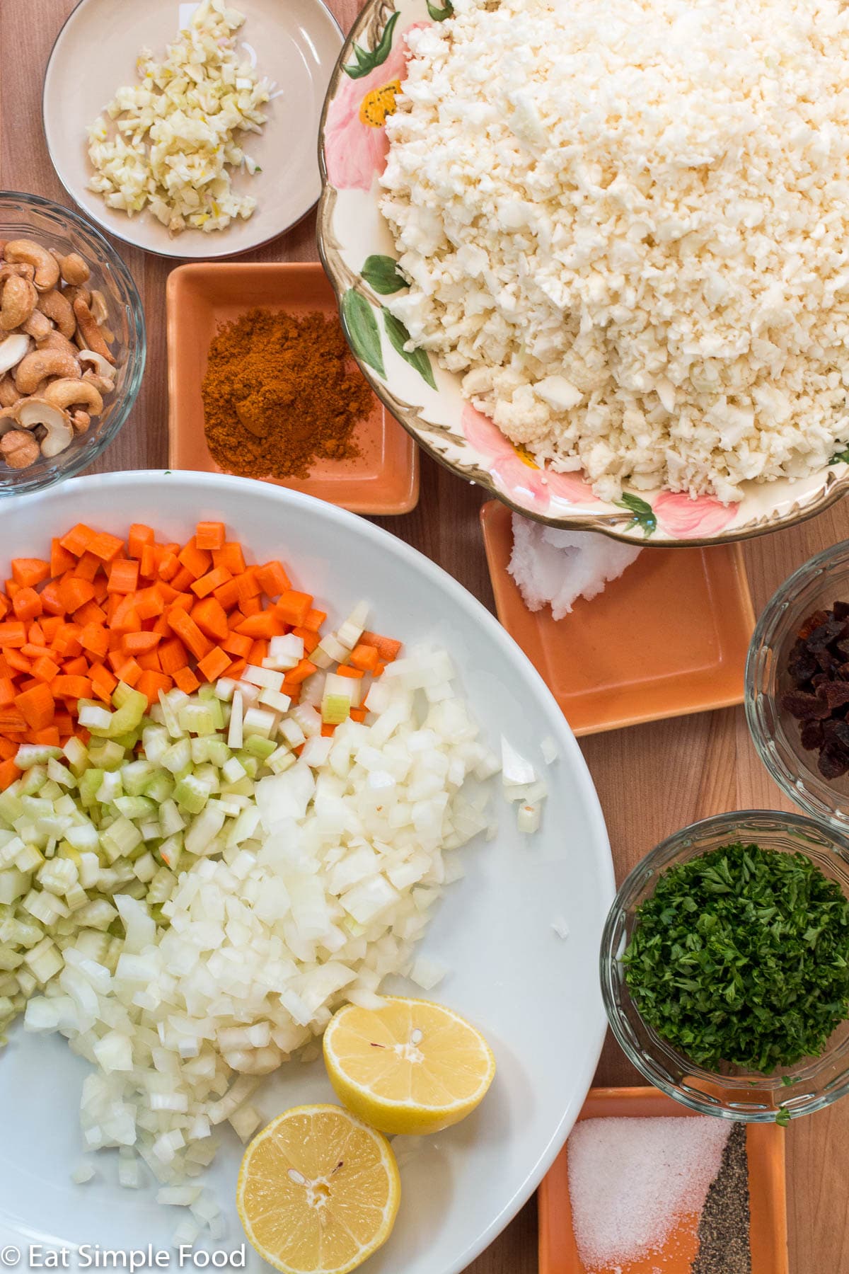 Ingredients on white plate, bowls, and small containers: diced carrots, celery, and onions, chopped parsley, cauliflower rice. garlic, halved lemons, salt, pepper, cashews, curry powder, and coconut oil.