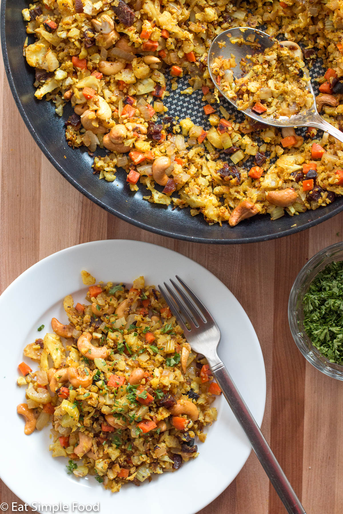 Top view of white plate of cashew yellow curry rice with raisins on a white plate with a fork. Pan with same thing on the side.