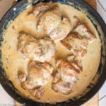 Top down view of cooked boneless skinless seared chicken thighs in a yellow cream mustard sauce in a dark pan.