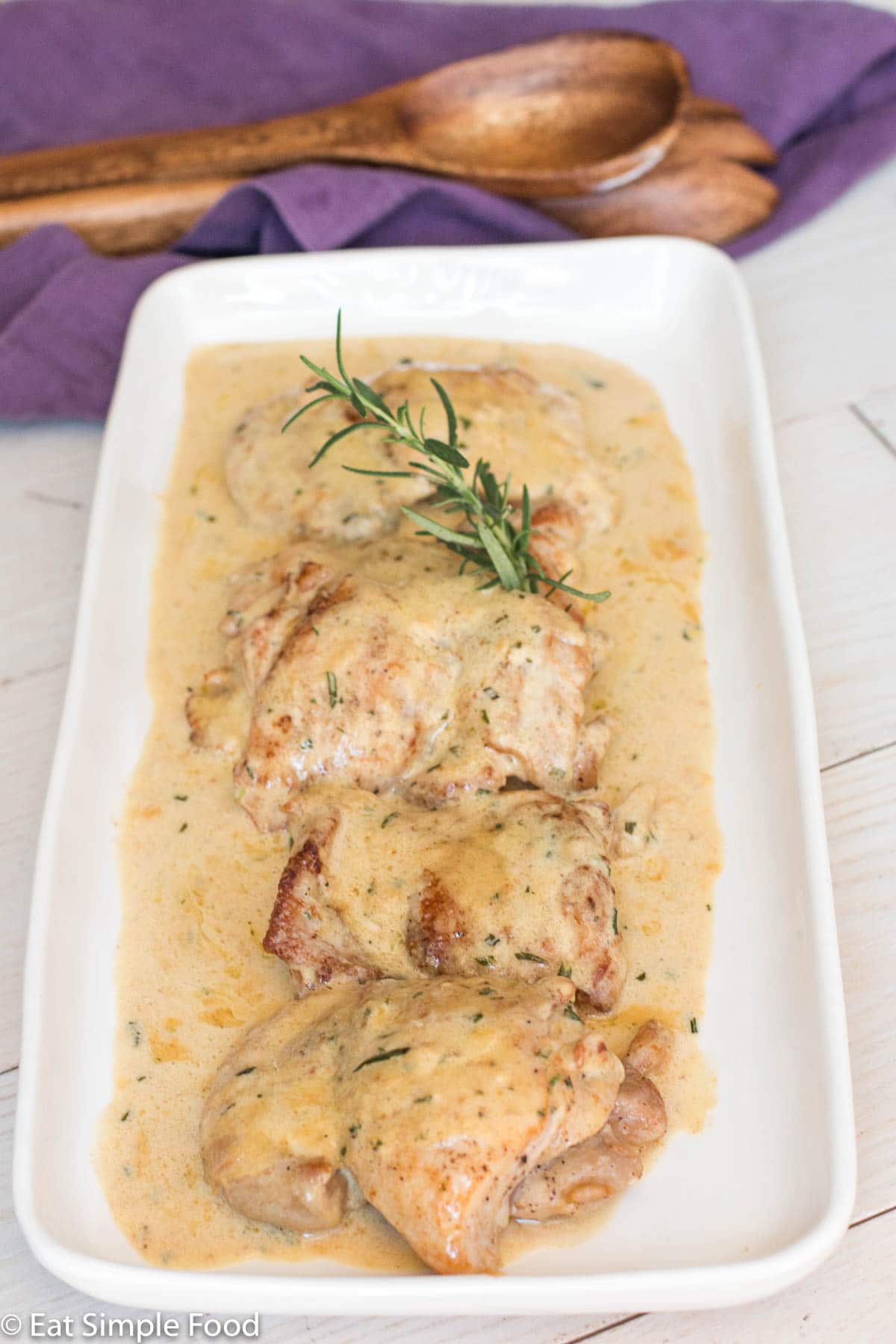 Side view of seared boneless chicken thighs on a white plate with a yellow mustard cream sauce with a sprig of fresh rosemary.
