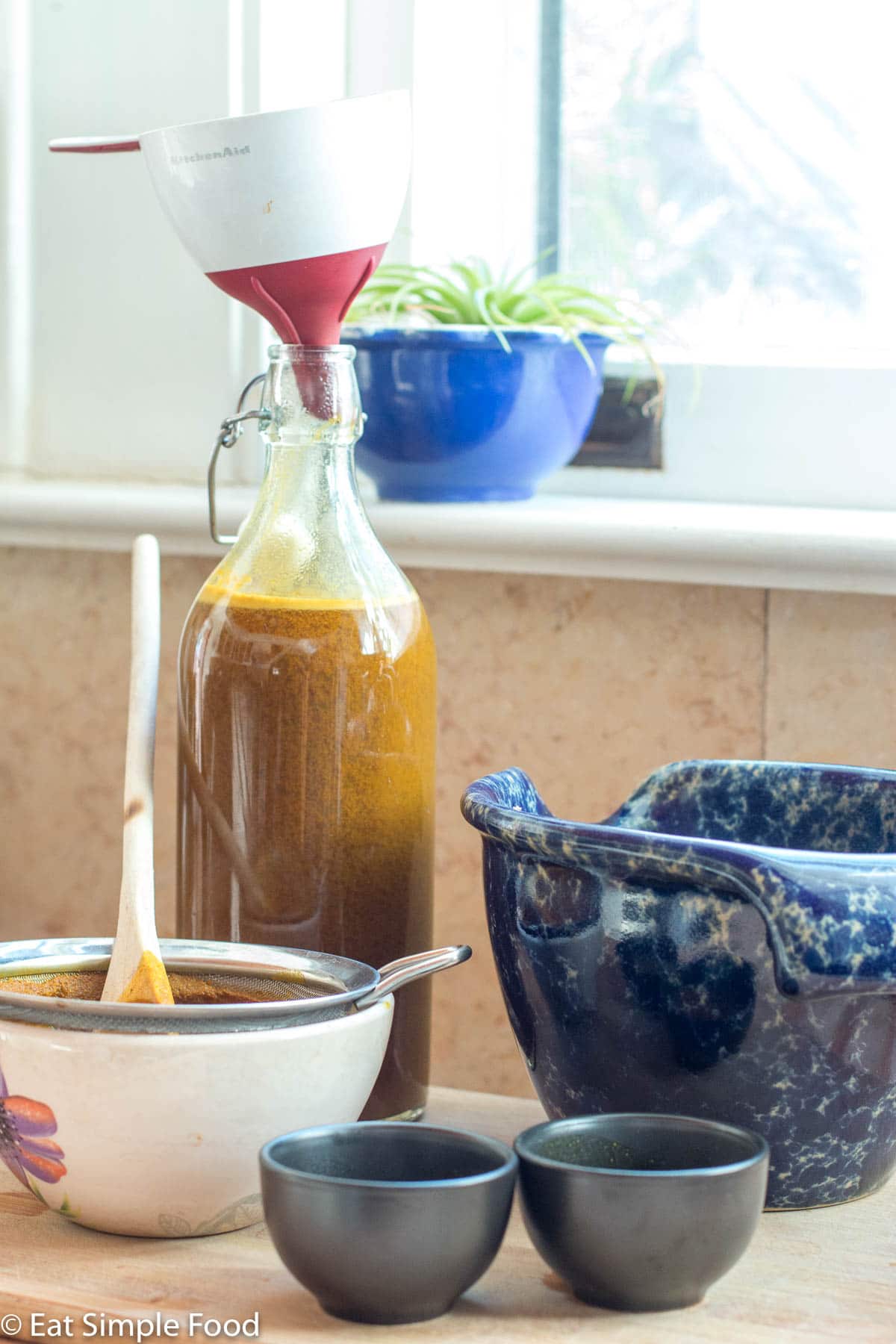 A liter size bottle with brown jamu liquid in it with a funnel on the top. White bowl in the front. Blue bowl on the side and two very small black drinking shot glasses in the front.
