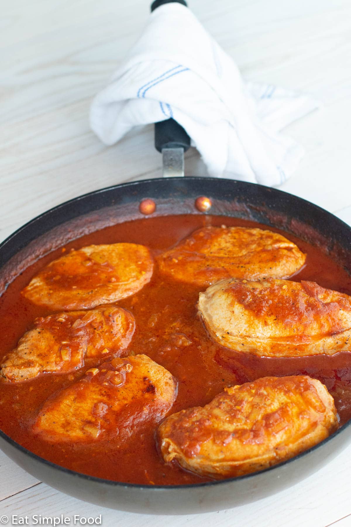 Pan of 6 boneless pork chops with red tomato gravy in a dark skillet on a white table. White napkin wrapped around the pan handle.