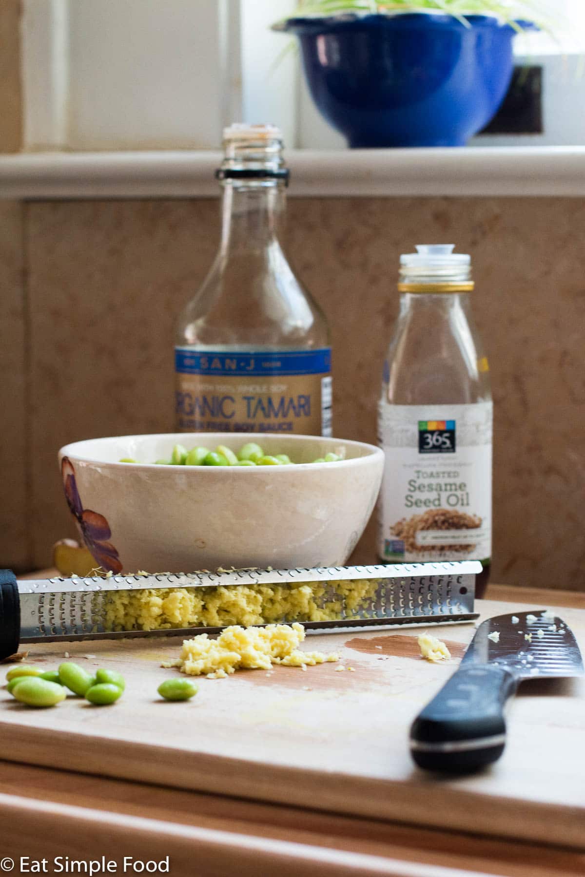 Ingredients on a wood cutting board: sesame oil bottle, soy sauce bottle, bowl of shelled edamame, minced ginger, chef's knife.