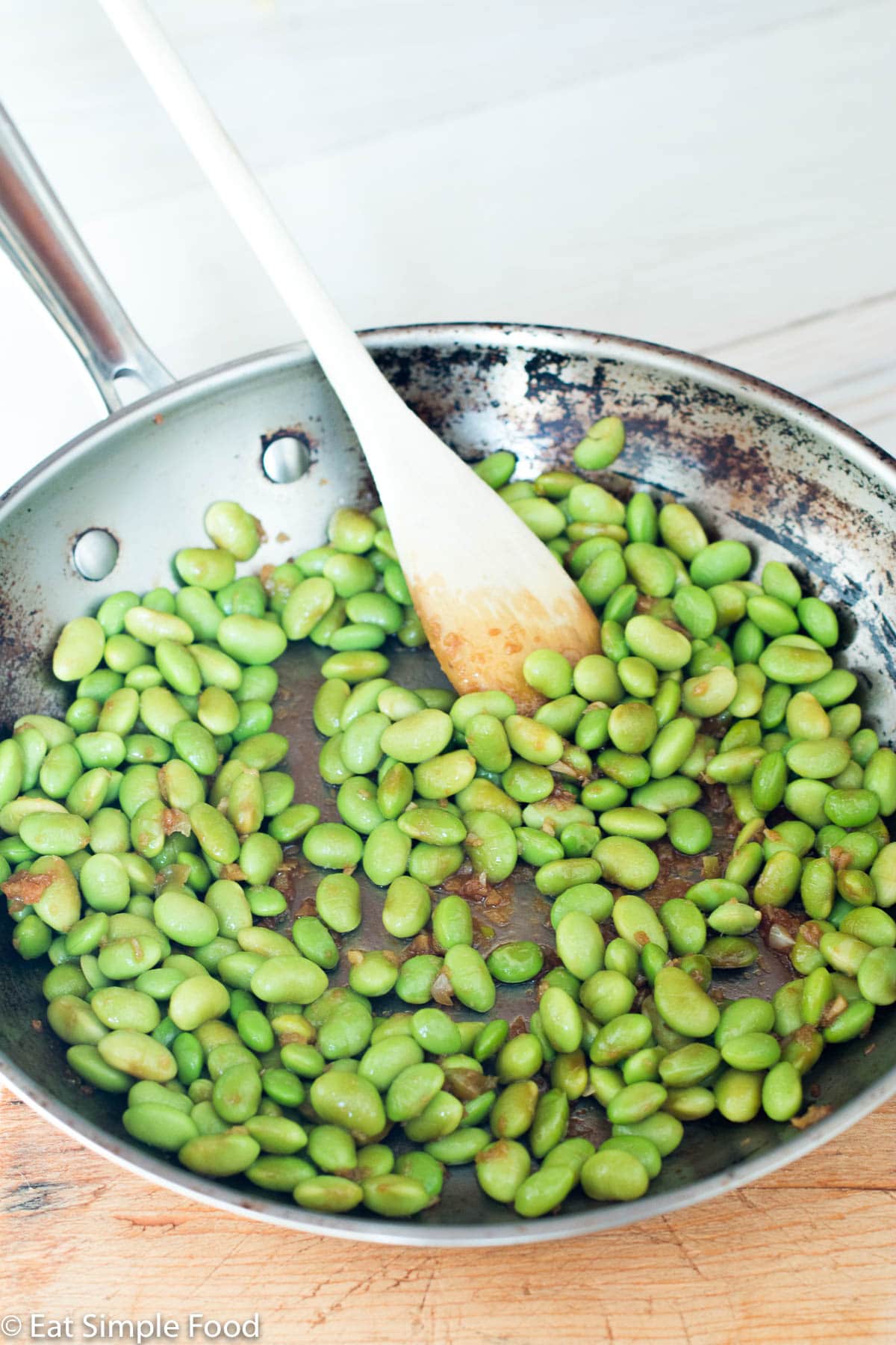 Stainless steel pan of shelled edamame cooked with garlic, ginger, and soy sauce. Wooden spoon hanging out the side.