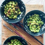 Top view of 3 black bowls of edamame with ginger and soy sauce on a wood cutting board with 3 sets of chopsticks.