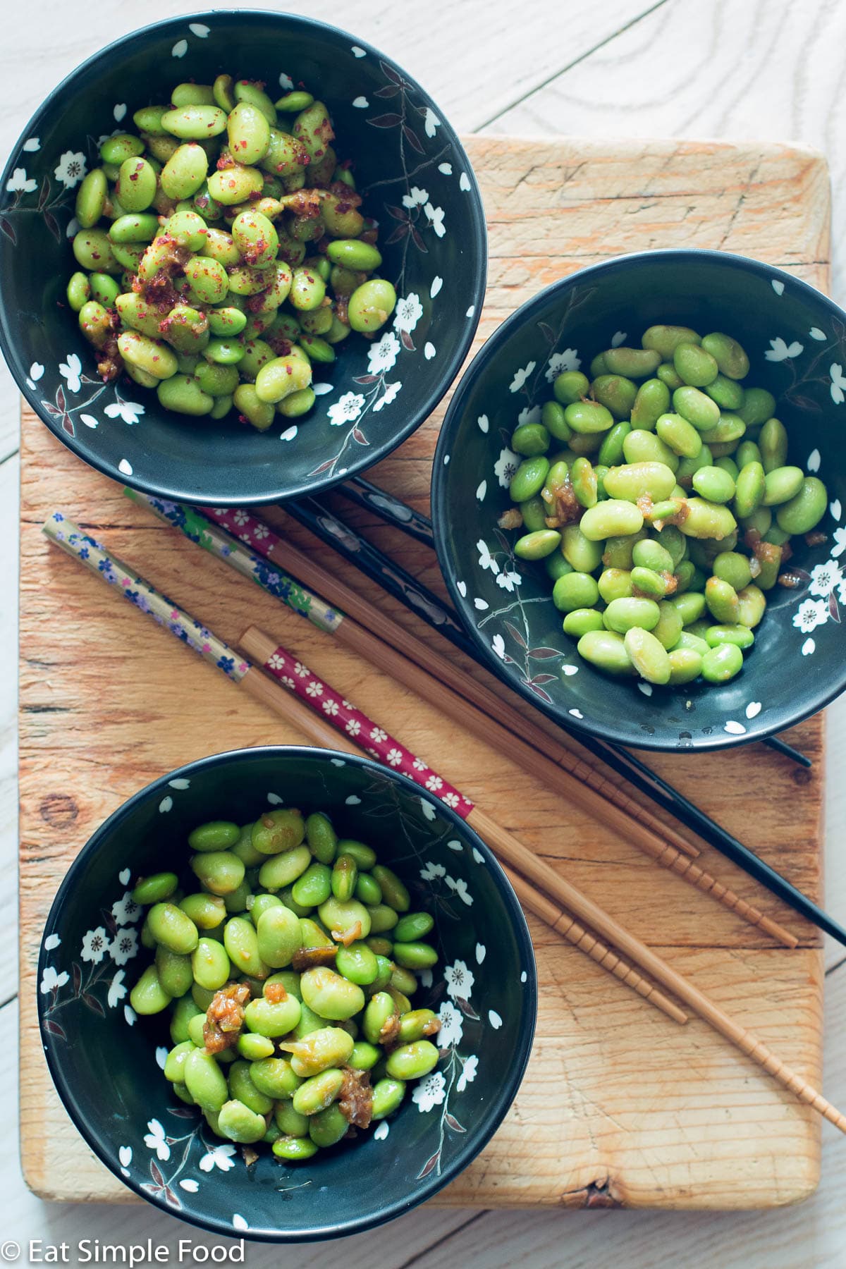 Top view of 3 black bowls of edamame with ginger and soy sauce on a wood cutting board with 3 sets of chopsticks.
