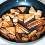 Pan fried and browned cooked tofu in a different shapes (wedges, cubes, slices, triangles) in a dark teflon pan. Side view.