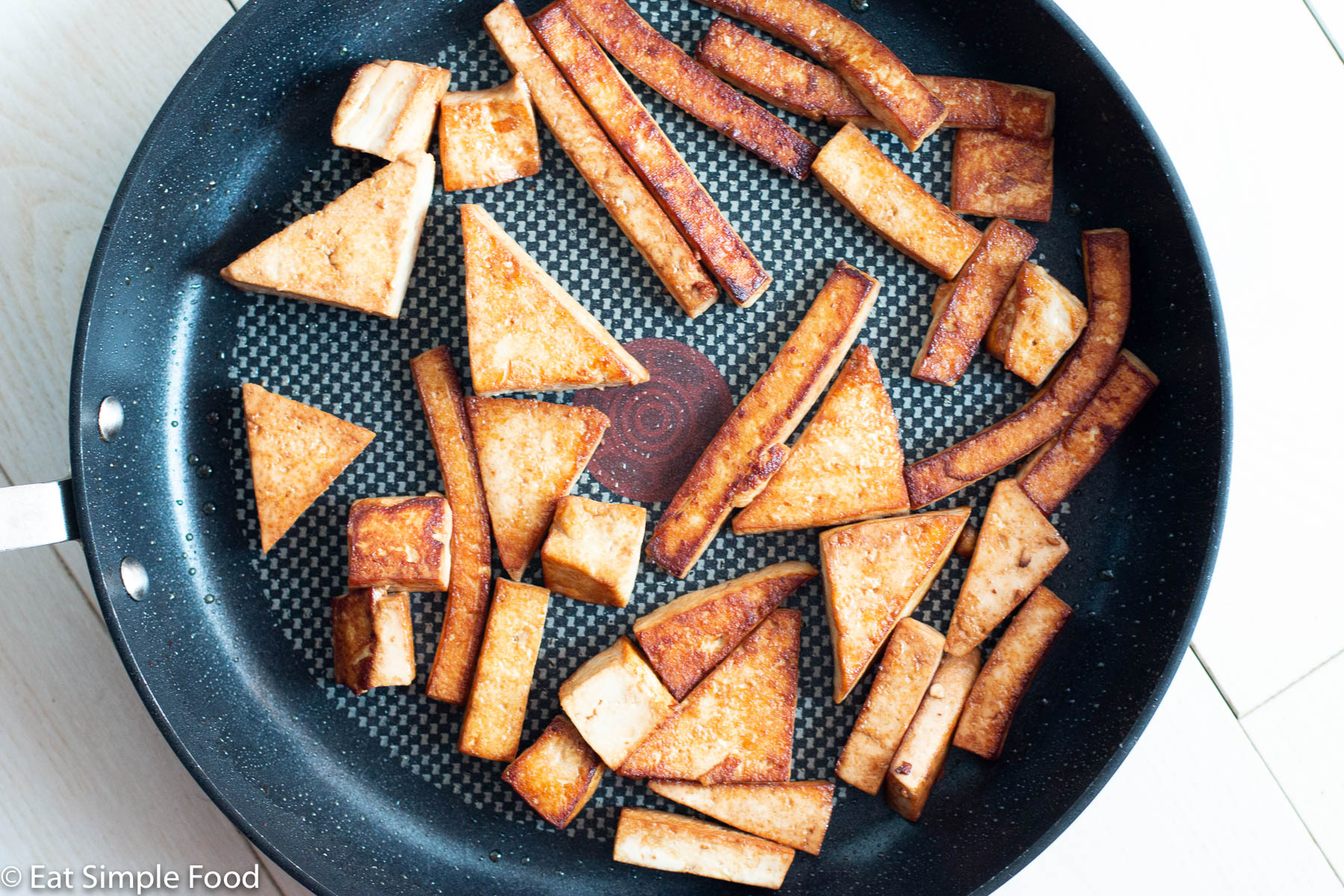 Pan fried and browned cooked tofu in a different shapes (wedges, cubes, slices, triangles) in a dark non stick pan. Top view.