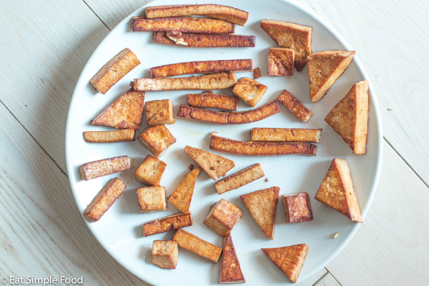 Pan fried and browned cooked tofu in a different shapes (wedges, cubes, slices, triangles) on a white plate on a white table.