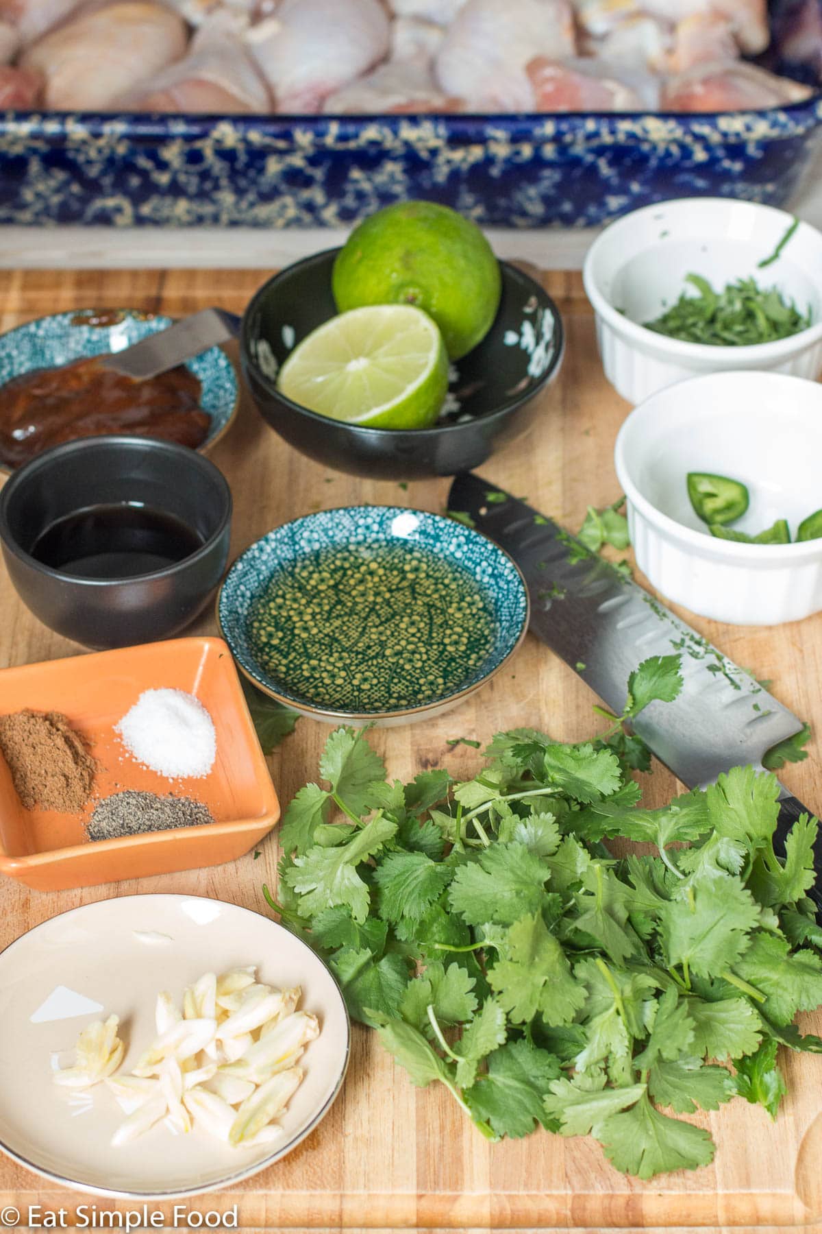 Raw ingredients on a wood cutting board: cilantro, garlic, spices, oil, lime cut in half, jalepeno, hoison sauce. Raw chicken in blue baking dish in background.