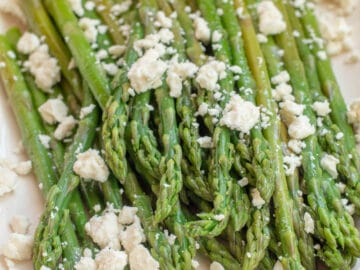 Rectangle white plate of cooked asparagus spears topped with crumbled feta cheese. Close up.