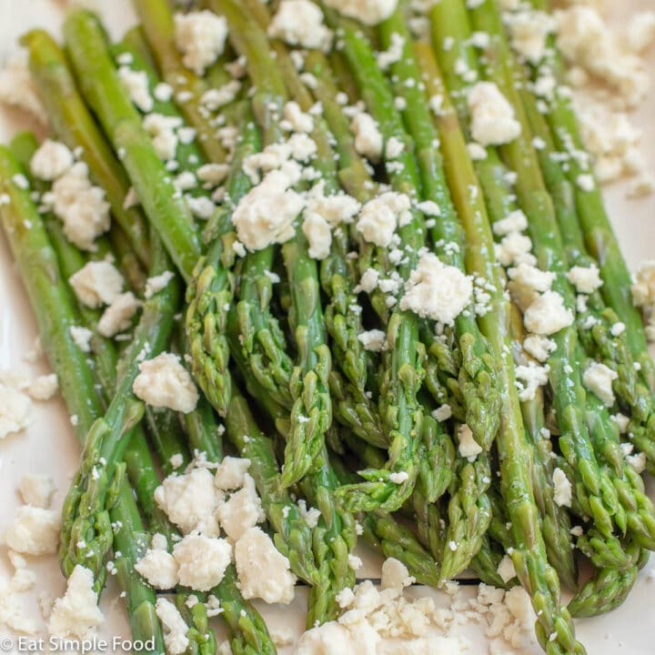 Rectangle white plate of cooked asparagus spears topped with crumbled feta cheese. Close up.
