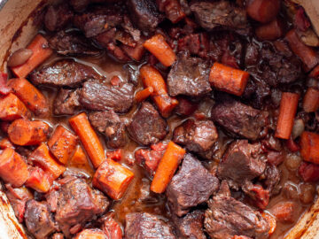 Top down view of red pot filled with cooked browned beef chunks and carrots in a brown gravy and tomato sauce.
