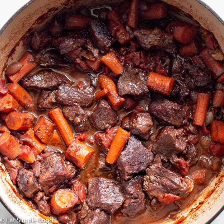 Top down view of red pot filled with cooked browned beef chunks and carrots in a brown gravy and tomato sauce.