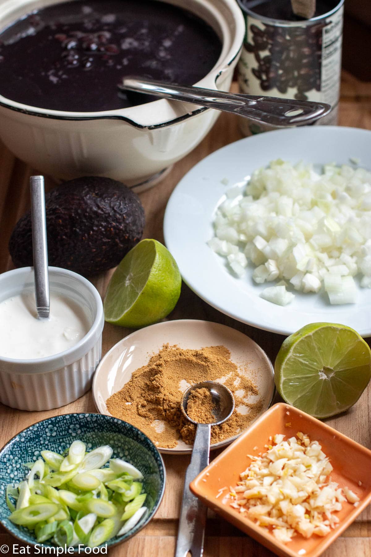 Ingredients: large bowl of black beans in liquid, bowl of sour cream, small bowl of raw chopped garlic, plate of diced onions, container of ground cumin with spoon, 2 lime halves, whole avocado, container of sliced green onions.