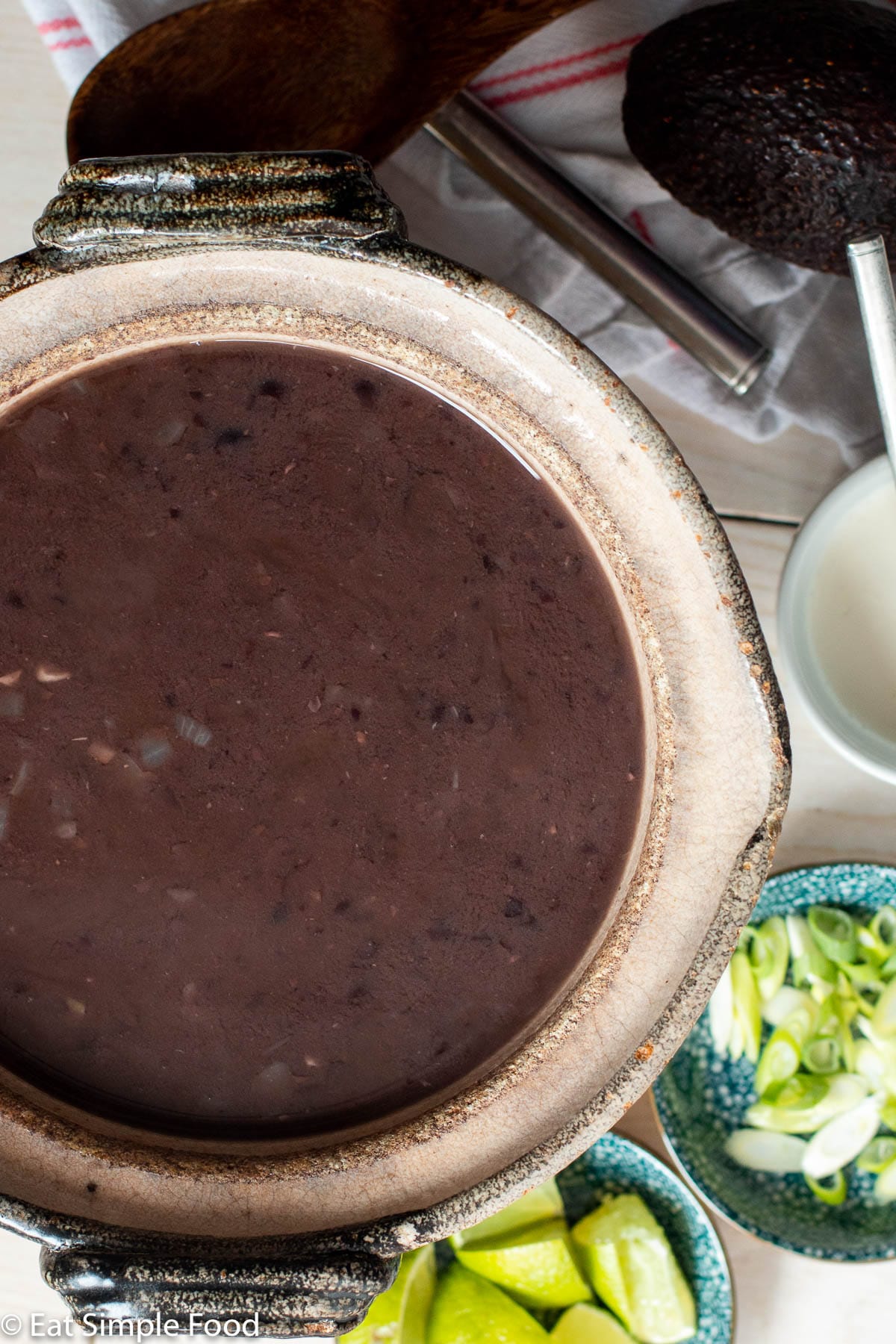 Top down view of bowl of dark black bean soup with lime wedges, sliced green onions, sour cream, and avocado in the background.