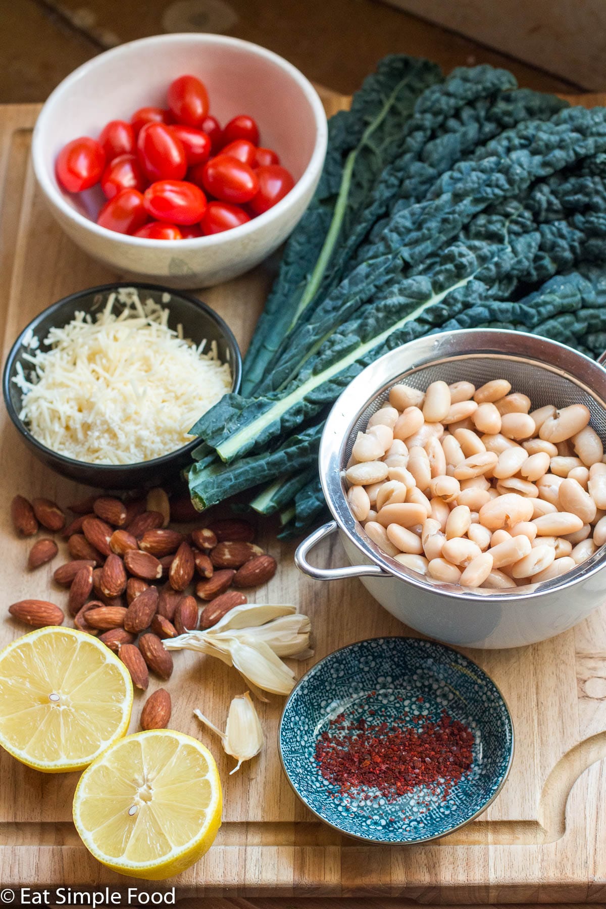 Ingredients on a wood cutting board. White beans in a strainer, bowl of cherry tomatoes, bowl of grated Parmesan cheese, bowl of chili pepper flakes, lemon cut in half, head of kale, whole almonds, and 4 garlic cloves. Side view.