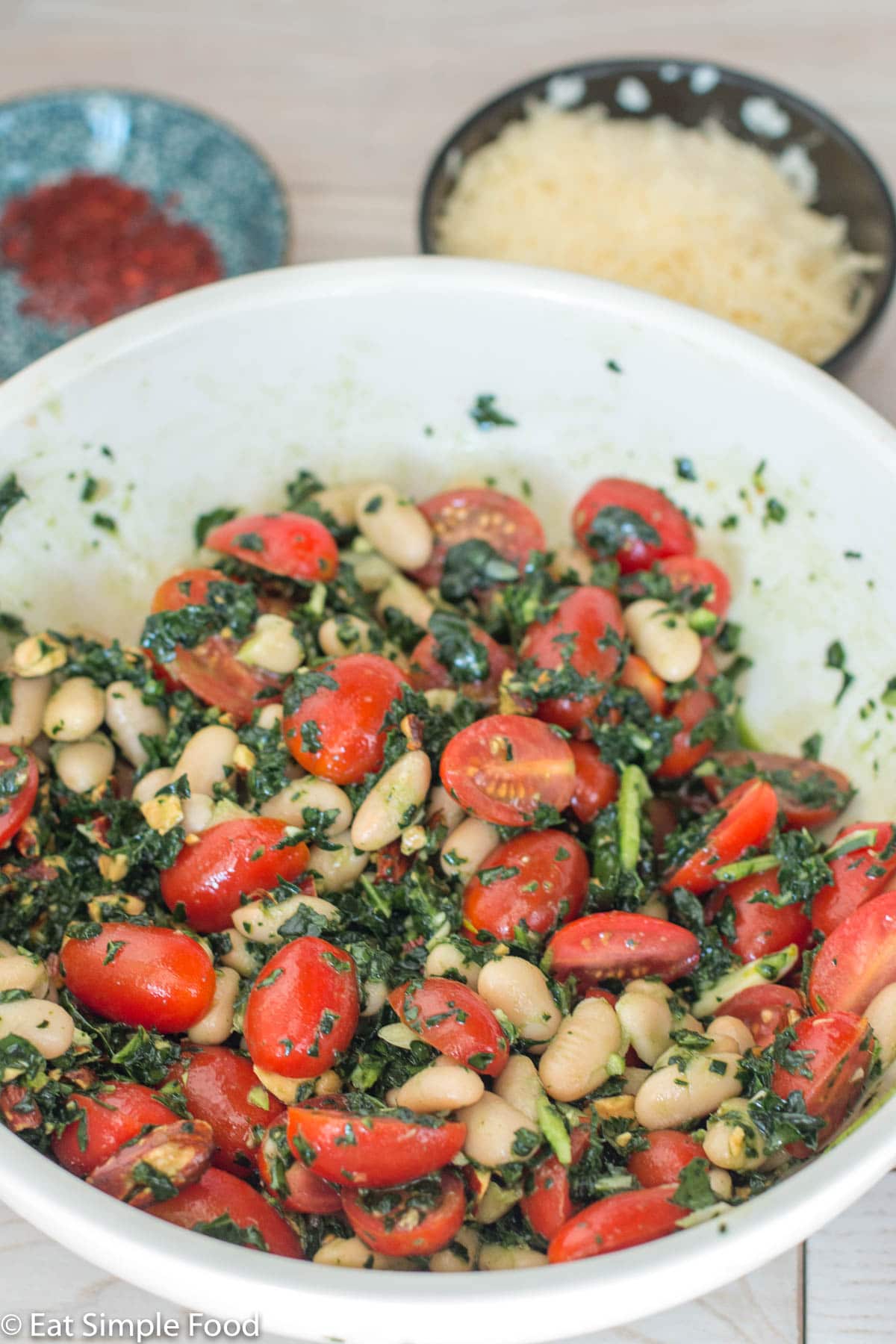 Side view of white beans and halved cherry tomatoes with Kale pesto in a white bowl. Bowl of chili flakes and grated Parmesan cheese in background.