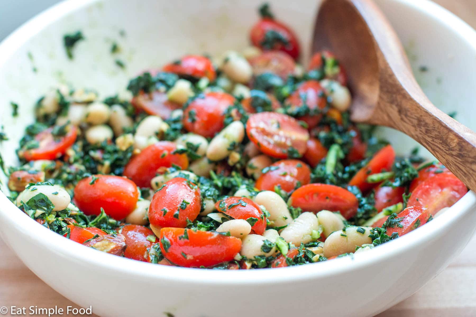 Side view of white beans and halved cherry tomatoes with Kale pesto in a white bowl with a wood spoon. Close up.