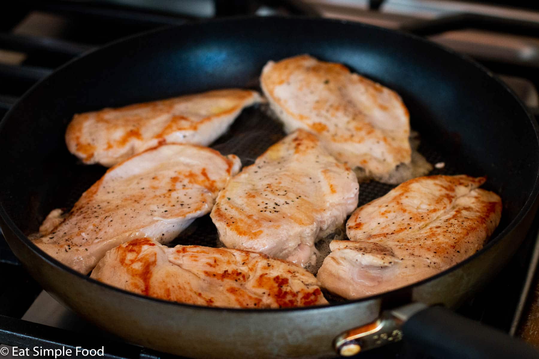 6 chicken cutlets cooking in a skillet on the stovetop. Cutlets are seared golden brown.