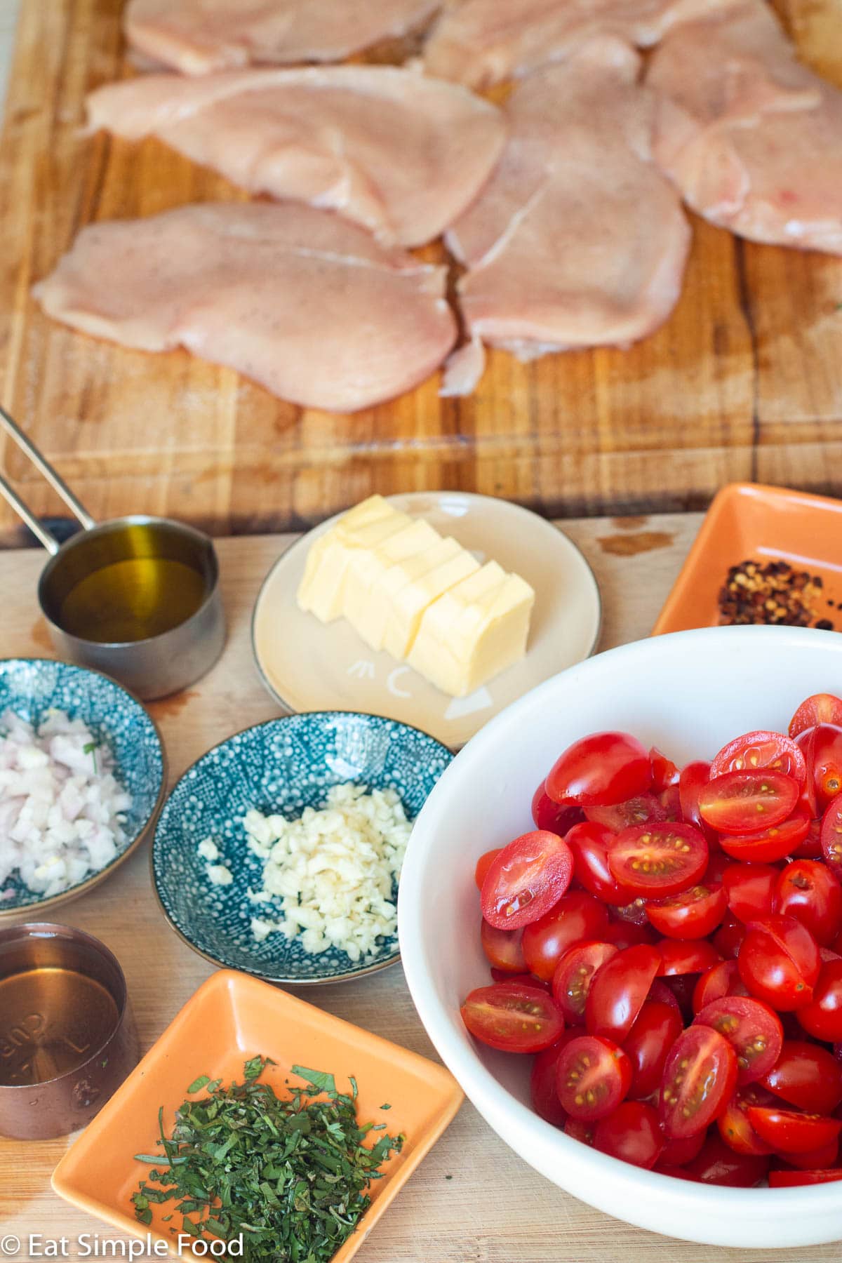 Ingredients: halved cherry tomatoes in a white bowl, little containers each of sliced butter, red chili flakes, minced shallots, minced garlic, chopped herbs and a wood cutting board with raw chicken cutlets on it.