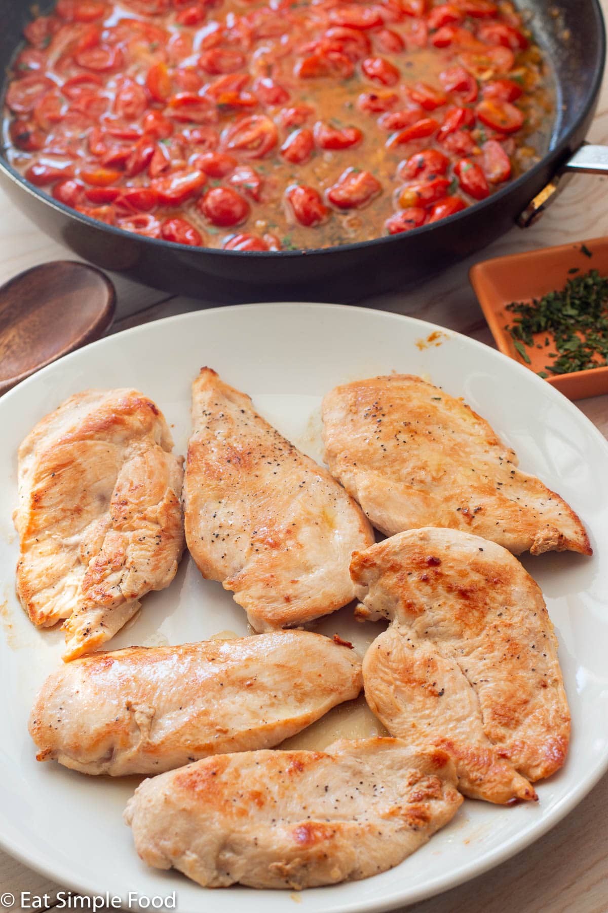 6 browned chicken cutlets on a white plate with a skillet of cooked cherry tomato sauce in background.