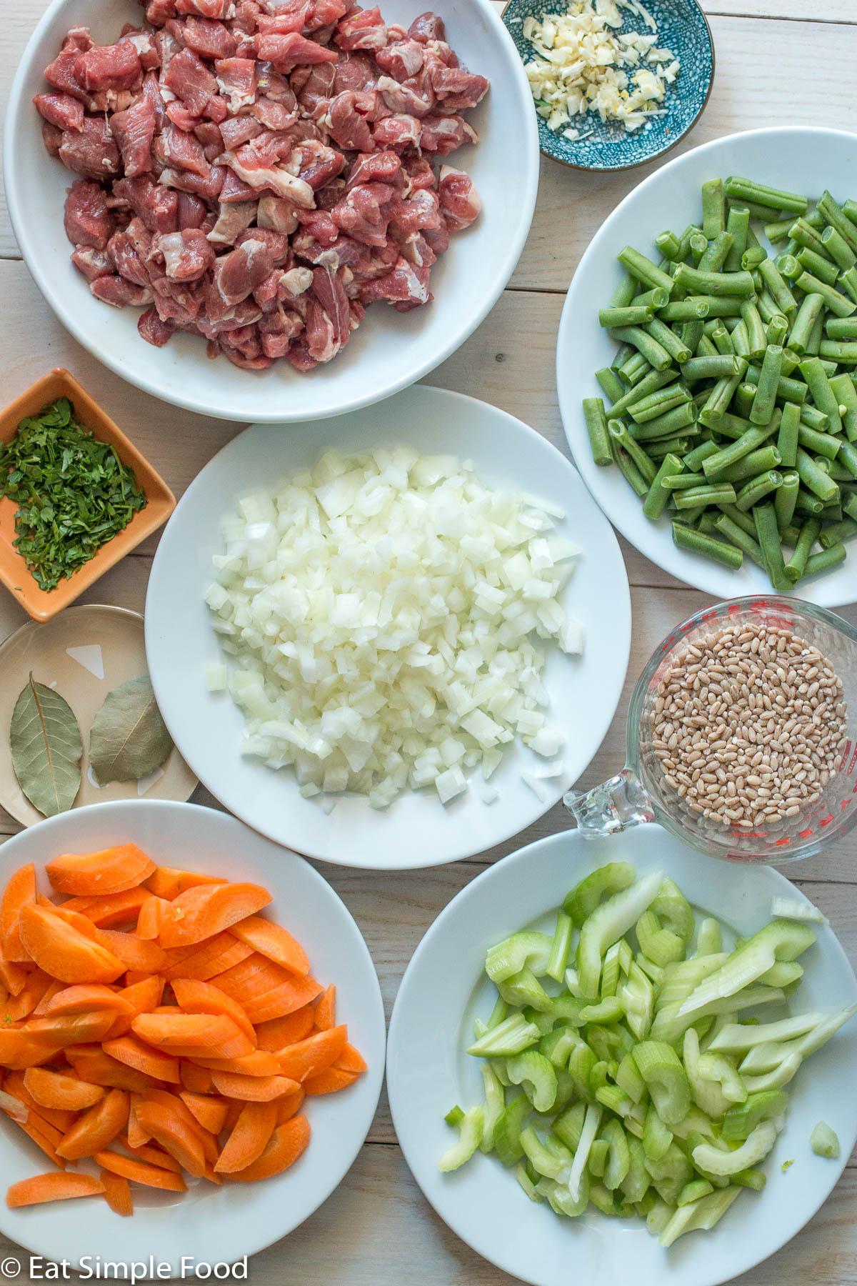 Top view of raw ingredients: plate of raw cubed lamb shoulder, plate of onions, small bowl of garlic, small bowl of chopped parsley, 2 bay leaves, measuring cup of pearl barley, plate of chopped green beans, plate of sliced carrots, plate of sliced celery.