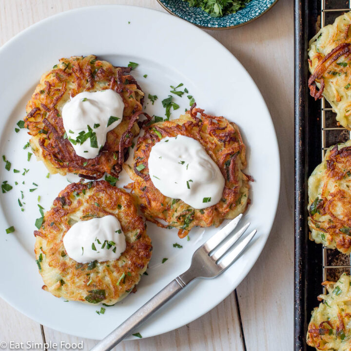 Top view (close up) of 3 potato latkes on white plate with sour cream and chopped parsley and a small fork. Baking sheet with 3 latkes in the background.