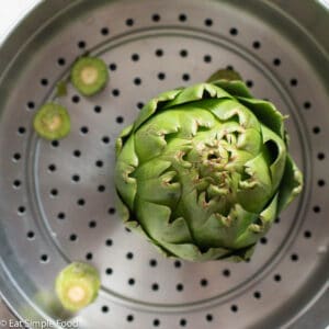 Top view of a raw artichoke sitting in a steaming basket of a large stainless steel pot. Top view.