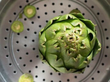 Top view of a raw artichoke sitting in a steaming basket of a large stainless steel pot. Top view.