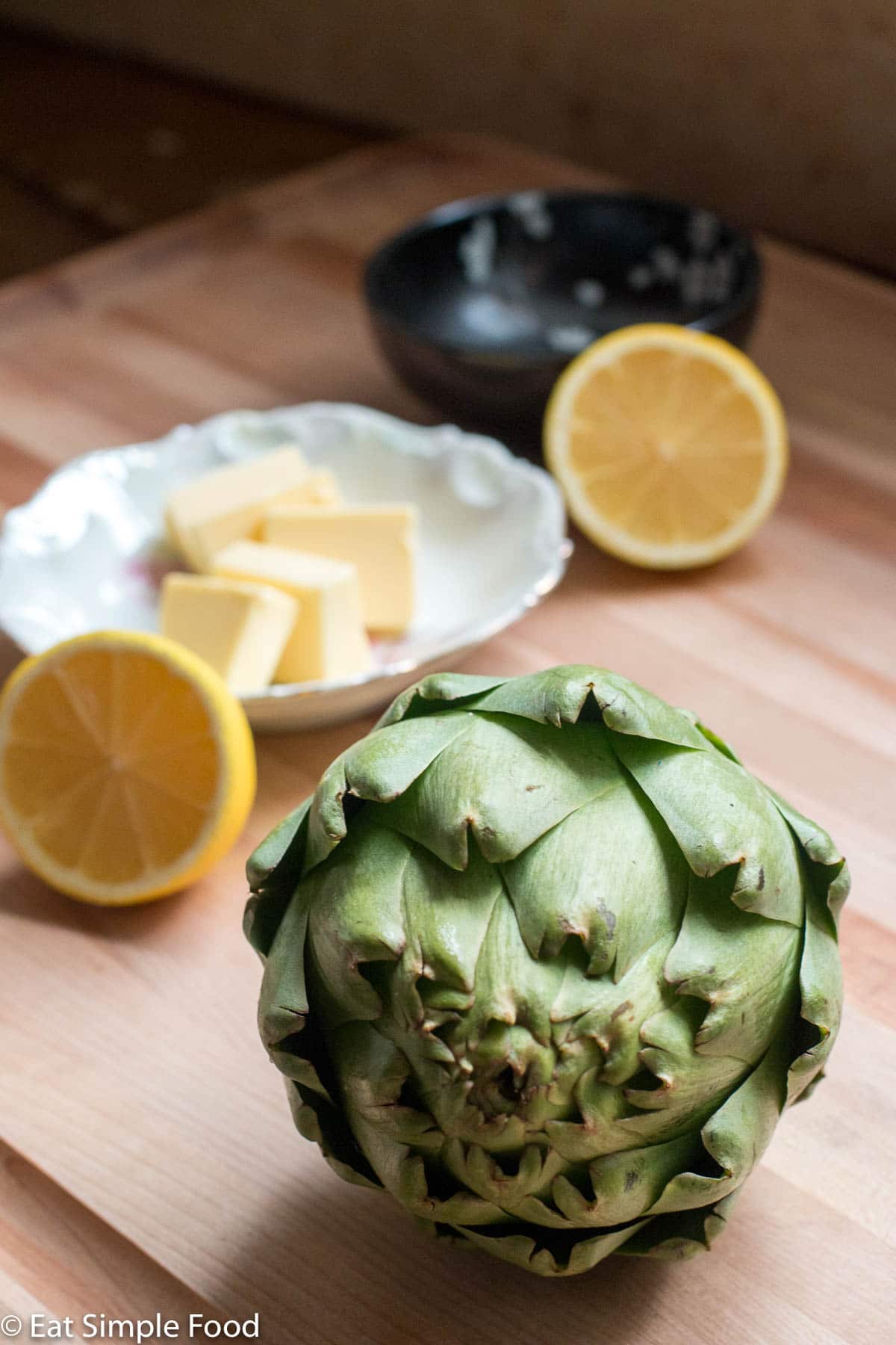 Raw artichoke on wood cutting board. 2 lemon halves and tabs of butter in a small white shallow bowl in the background.
