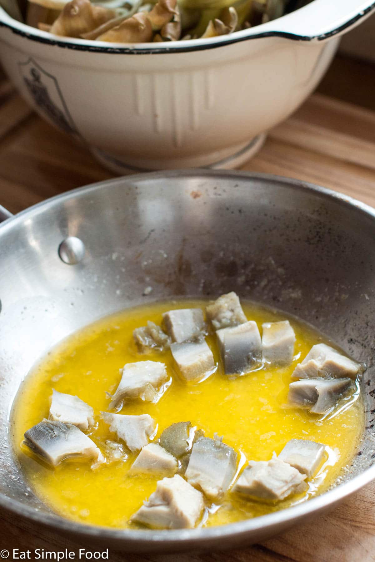 Cooked diced artichoke hearts in a stainless steel pan full of bright yellow butter.