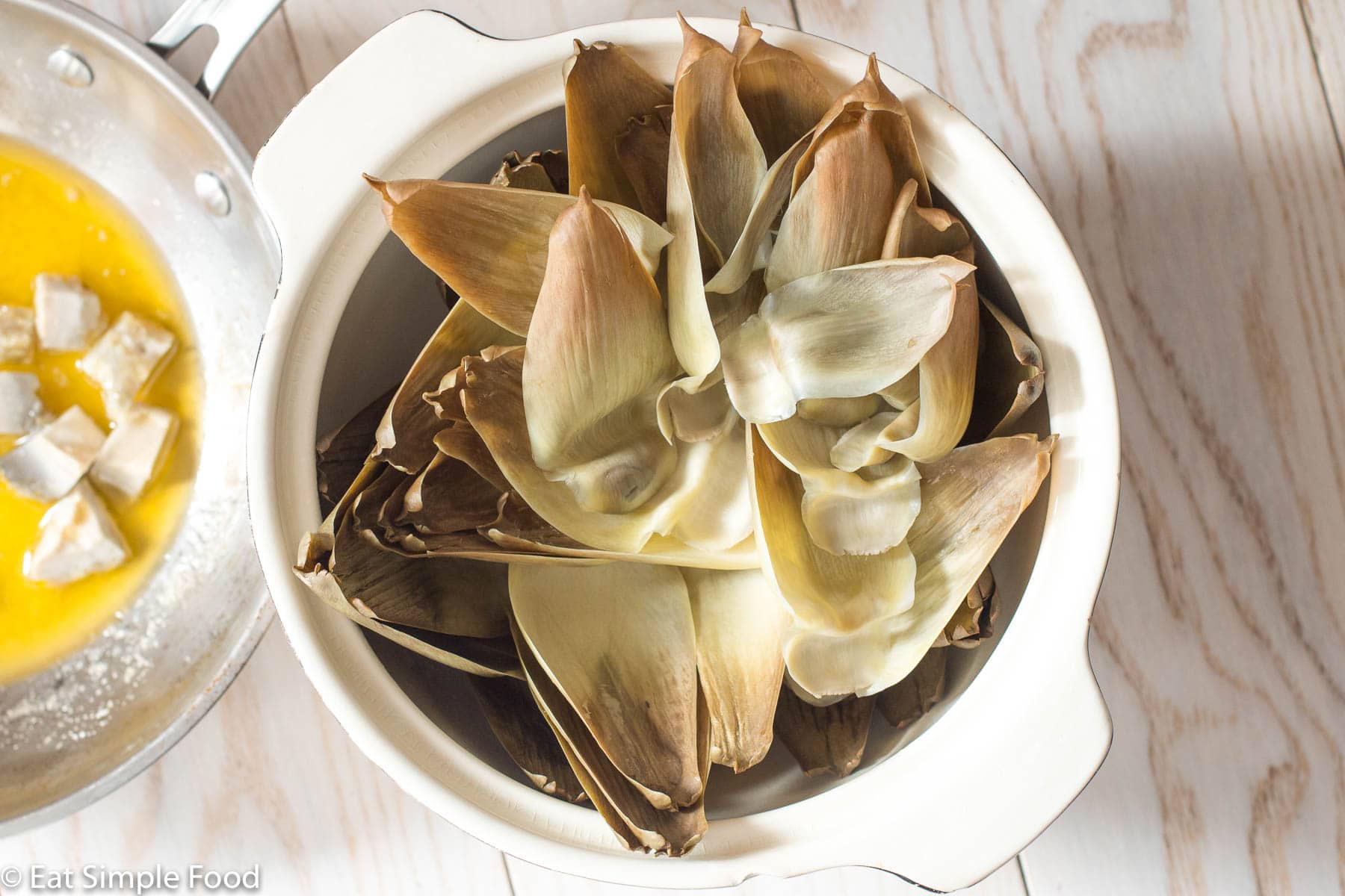Edible cooked artichoke leaves in a white bowl. Small saucepan of diced artichoke hearts marinating in butter on the side. Top view.