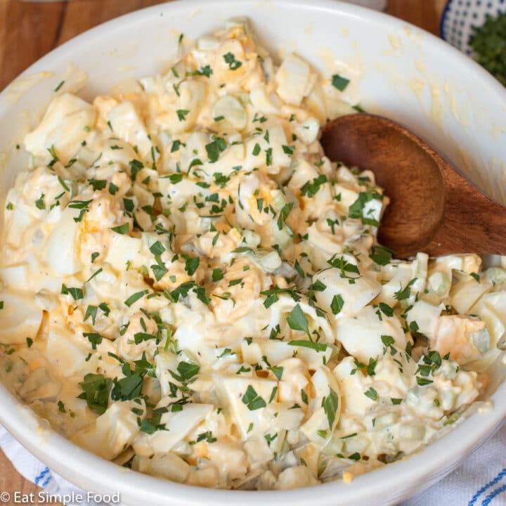 Egg Salad mixed with mayonnaise and topped with chopped parsley in a white bowl with a wood spoon with a white napkin underneath the bowl. Very Close up.