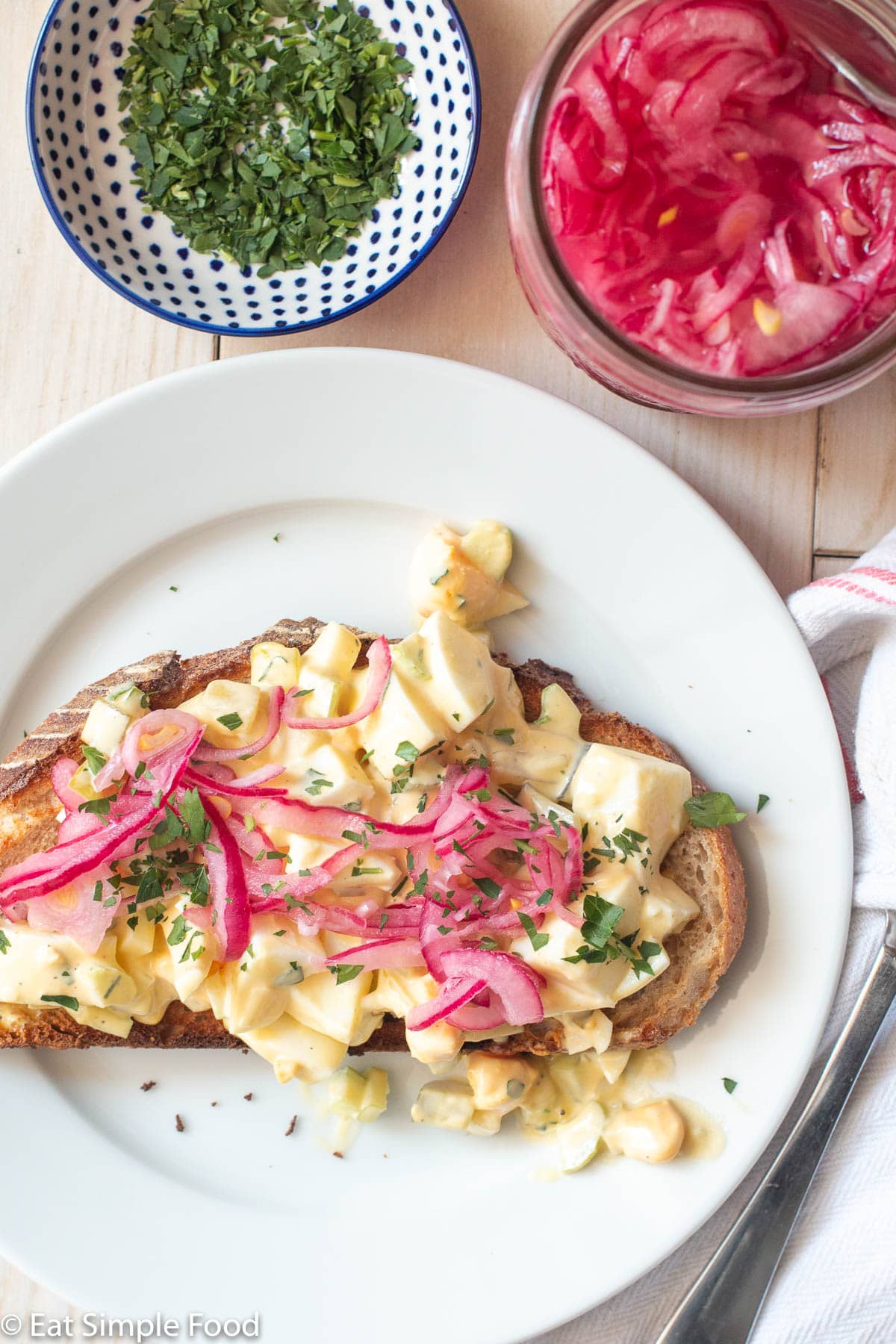 Top view of toast topped with a chunky egg salad and pickled red onions on a white plate with napkin and fork on the side and small bowl of chopped parsley and jar of pickled red onions.