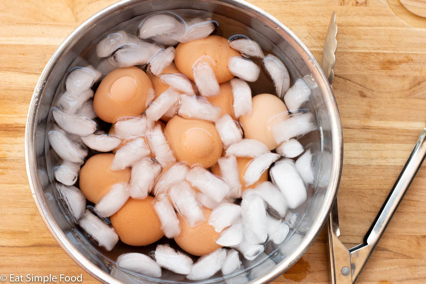 12 eggs in a stainless steel bowl filled with ice and water. Tongs on the side. On a wood cutting board. Top view.