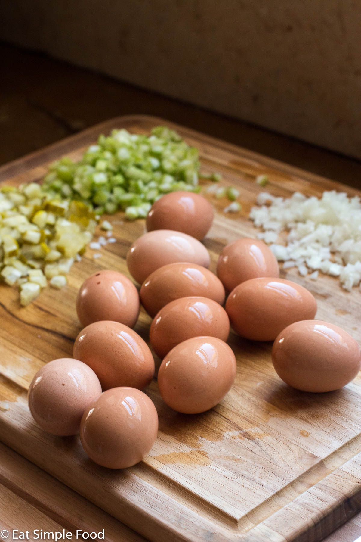 12 boiled eggs on a wood cutting board with chopped pickles, onions, and celery in the background.