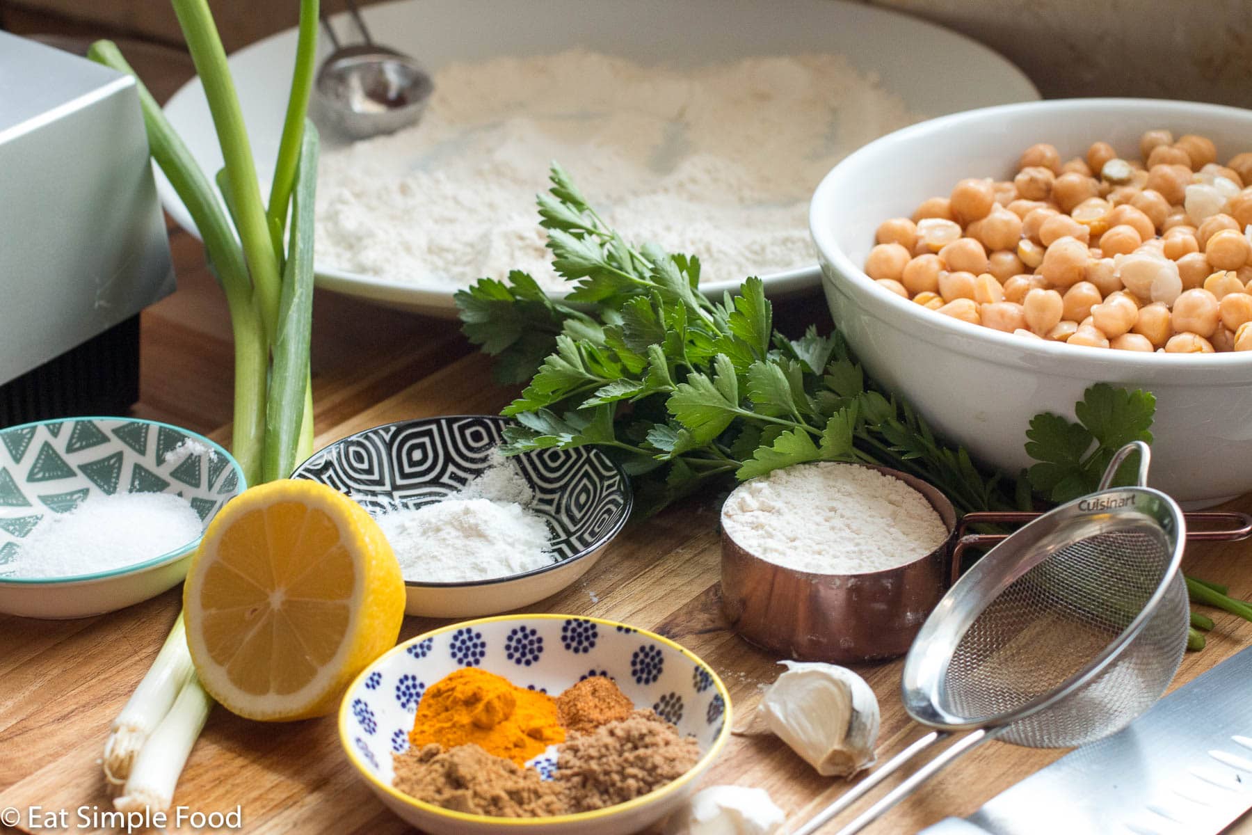 Ingredients on a wood cutting board: container of salt, baking powder, spices, measuring cup of flour, 2 cloves of garlic, bunch of parsley, white bowl of chickpeas, plate of flour, 2 green onions, ½ lemon. Side view.